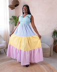 A woman wearing a sleeveless multi-colored flared maxi dress with blue, yellow, and pink tiers, standing in a cosy modern room with natural lighting.