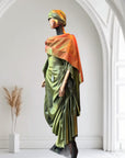 Olive green satin gown with ruched side draping, paired with a bronze-toned shawl and matching headwrap, displayed on a mannequin in an arched interior.
