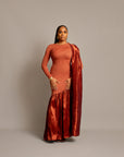 A woman wearing a rust sari dress with an asymmetrical neckline, gathered satin hem, and draped fabric over one shoulder.