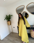 A woman wearing a bright yellow satin slanted dress with long puff sleeves and a high-low hemline, posing indoors near a mirror and window.