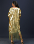A woman wearing a shimmering gold kaftan dress with long green fringes, standing against a dark background.
