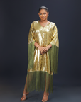 A woman wearing a shimmering gold kaftan dress with long green fringes, standing against a dark background.
