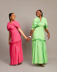 Two women wearing the Ine maxi dress in pink and green, featuring a draped V neckline, gathered waist detailing, and ruched hem accents.
