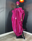 Pink off-shoulder boubou dress with black grid pattern, paired with a matching headwrap, displayed on a mannequin against a black paneled wall.
