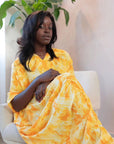 Woman wearing a yellow and orange tie-dye draped maxi dress with front gathers, standing indoors.
