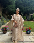 Woman in a flowing champagne gold Demure Boubou with matching headwrap, standing in a garden walkway with flowers.
