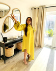 A woman wearing a bright yellow satin slanted dress with long puff sleeves and a high-low hemline, posing indoors near a mirror and window.
