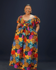 A woman wearing a vibrant floral maxi dress with puff sleeves, a front bow detail, and side pockets against a dark background.
