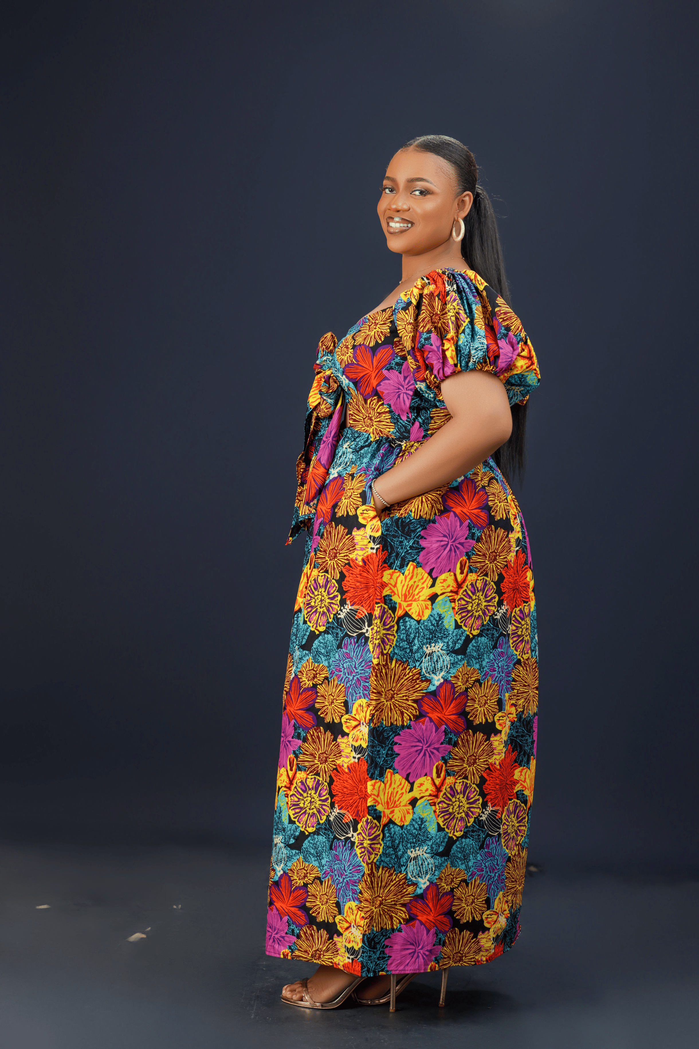 A woman wearing a vibrant floral maxi dress with puff sleeves, a front bow detail, and side pockets against a dark background.
