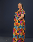 A woman wearing a vibrant floral maxi dress with puff sleeves, a front bow detail, and side pockets against a dark background.
