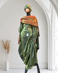 Olive green satin gown with ruched side draping, paired with a bronze-toned shawl and matching headwrap, displayed on a mannequin in an arched interior.
